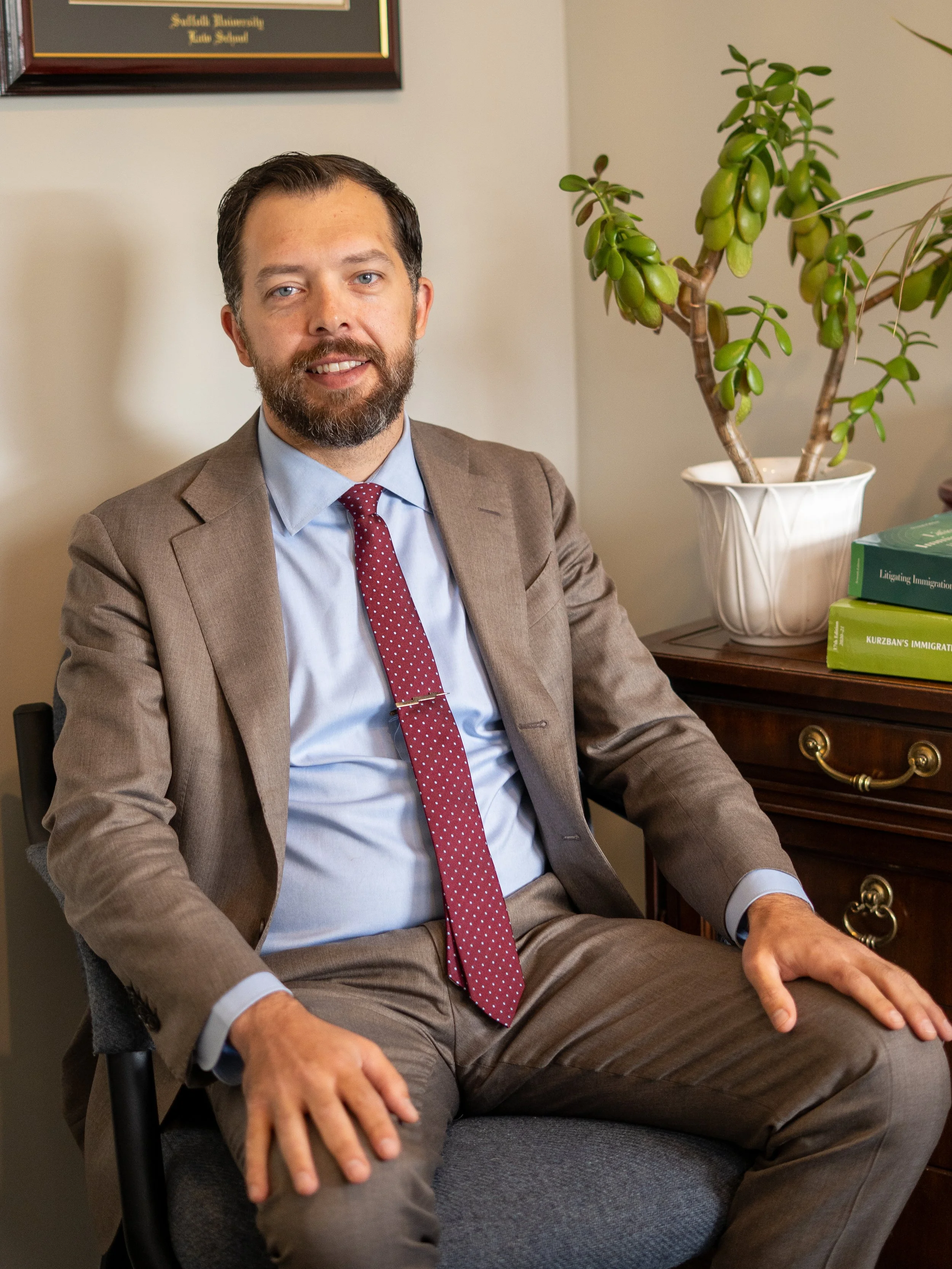 A man with a beard and blue eyes sitting in an office, wearing a brown suit, light blue shirt, and a red polka dot tie. Behind him is a dresser with a potted succulent plant and books about immigration.
