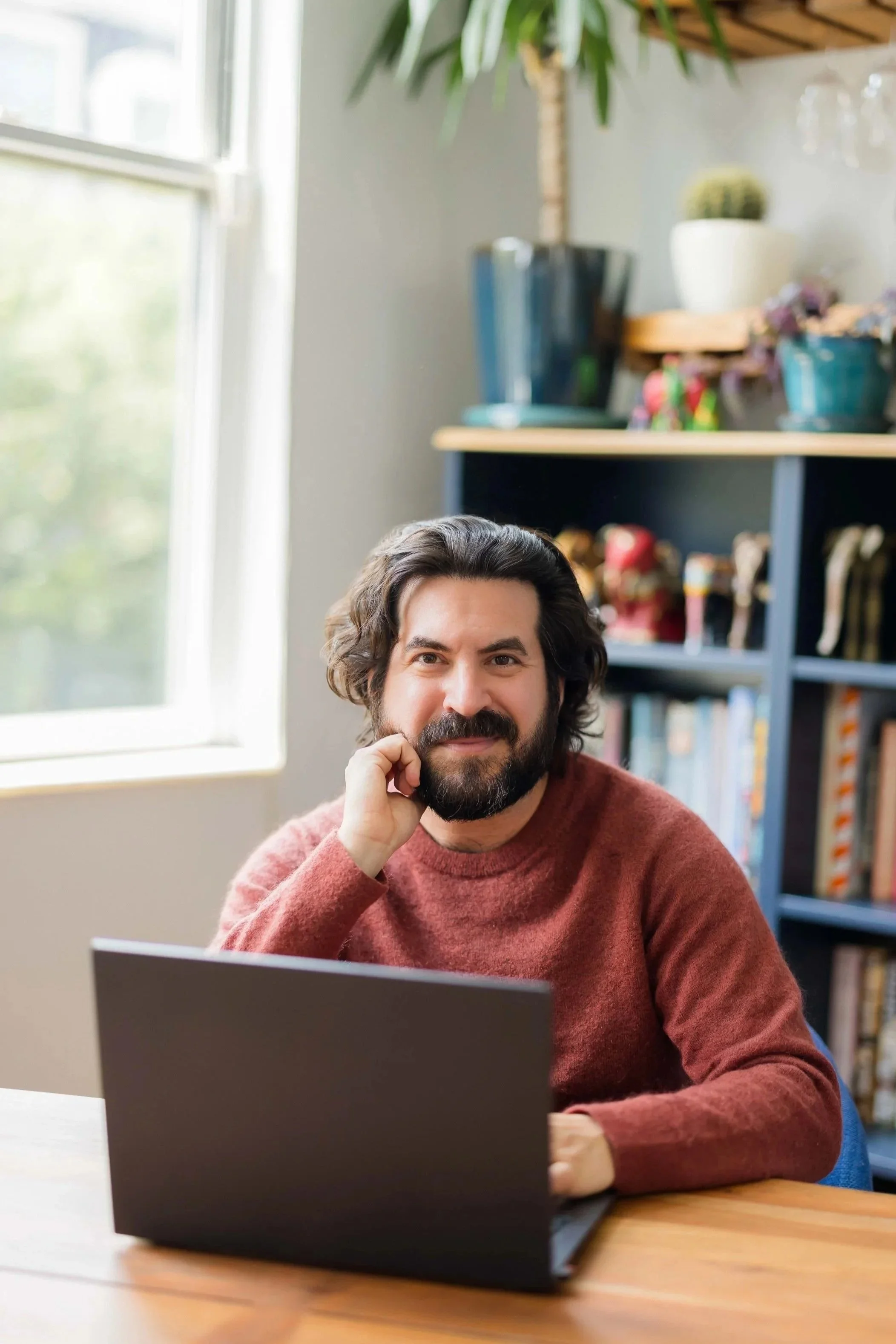 A man with dark, wavy hair and a beard, wearing a maroon sweater, sitting at a wooden desk in front of a laptop, smiling and looking at the camera, with a bookshelf and a window in the background.