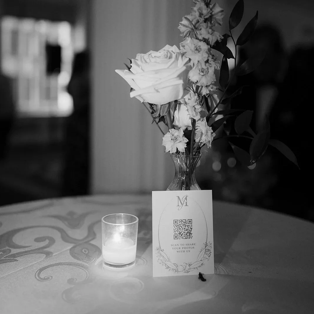 A black-and-white photo of a flower vase with roses and other flowers on a table. A candle and a branded card with a QR code are also on the table.