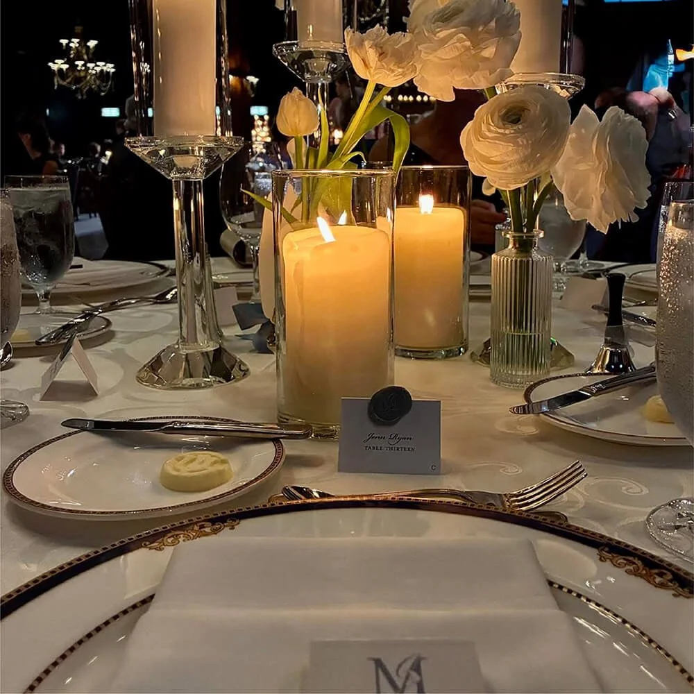 A formal dining table decorated with lit candles, white flowers in vases, and elegant tableware, set for a wedding. The place card has a branded wax seal and there is a matchbox in the foreground.