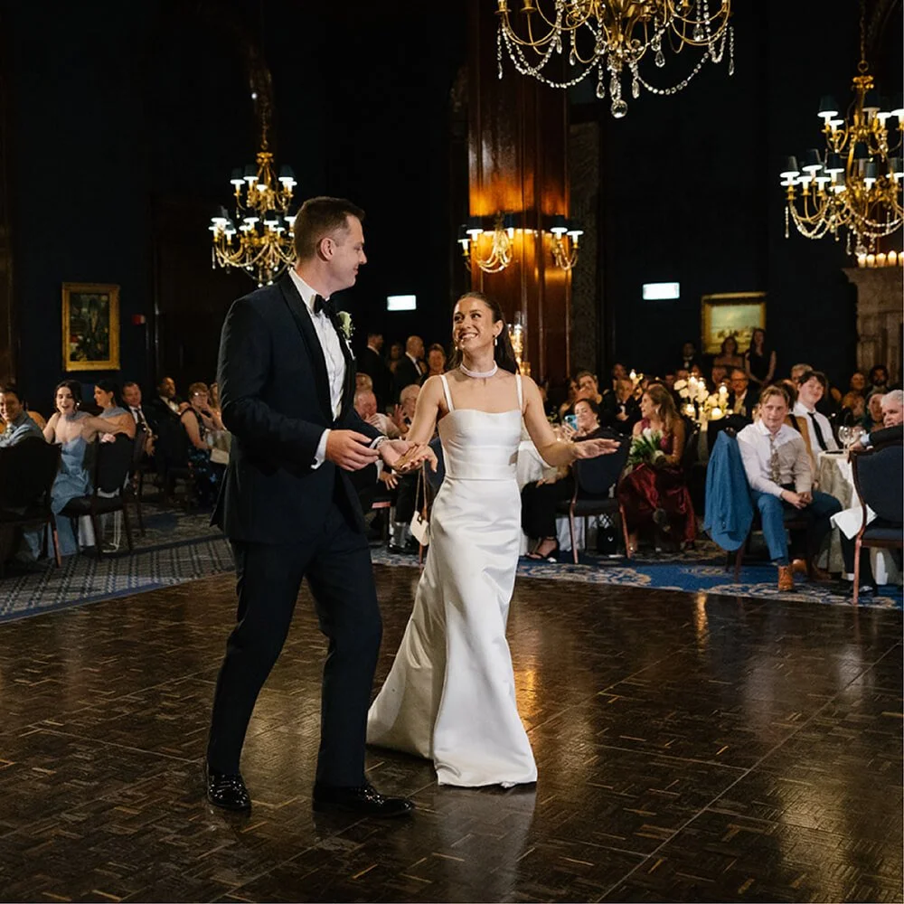 Bride and groom dancing at their wedding reception in a grand hall with chandeliers, guests seated at tables watching.