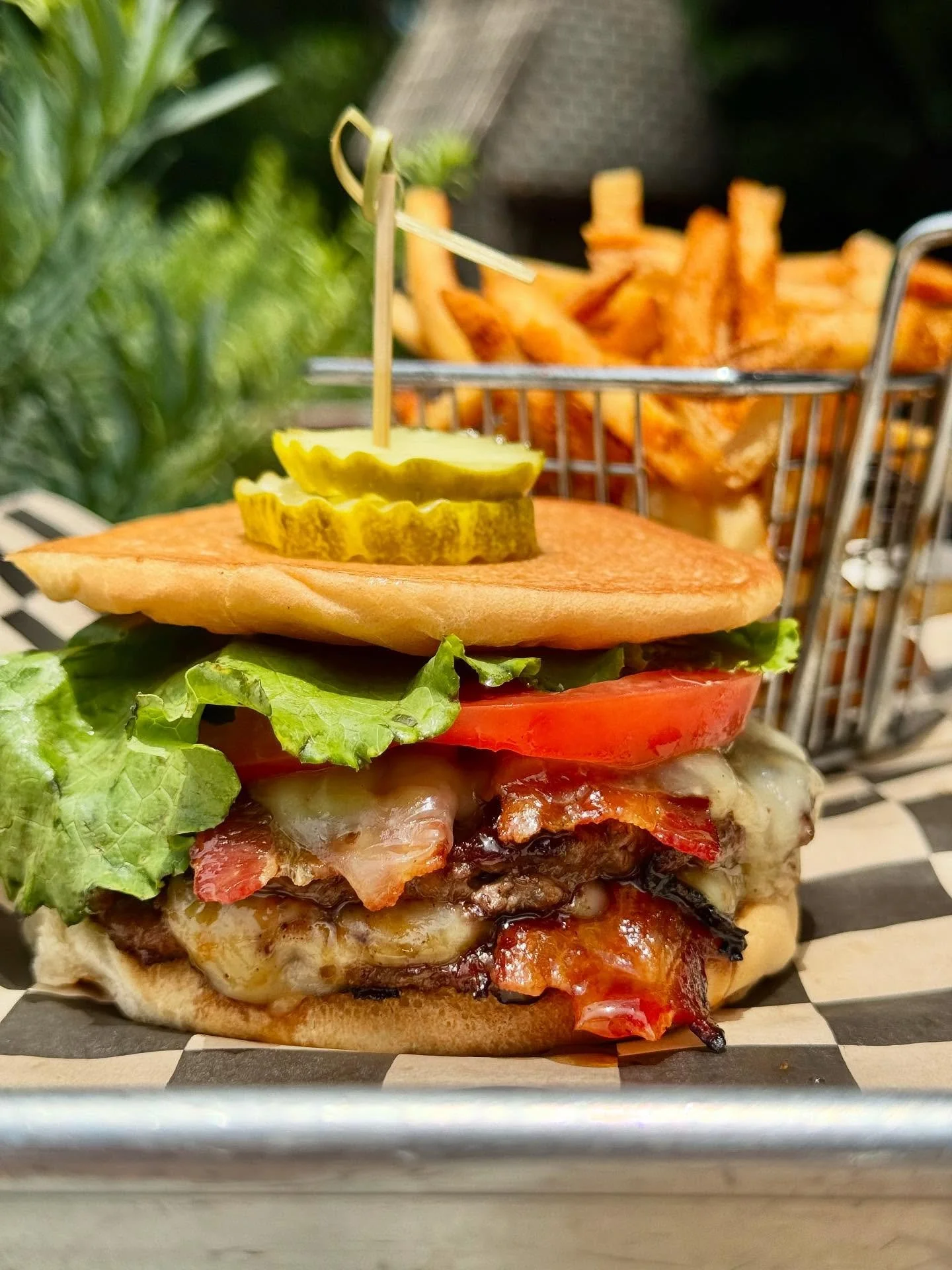 Close-up of a hamburger with lettuce, tomato, cheese, bacon, and pickles, served with French fries in a metal basket.