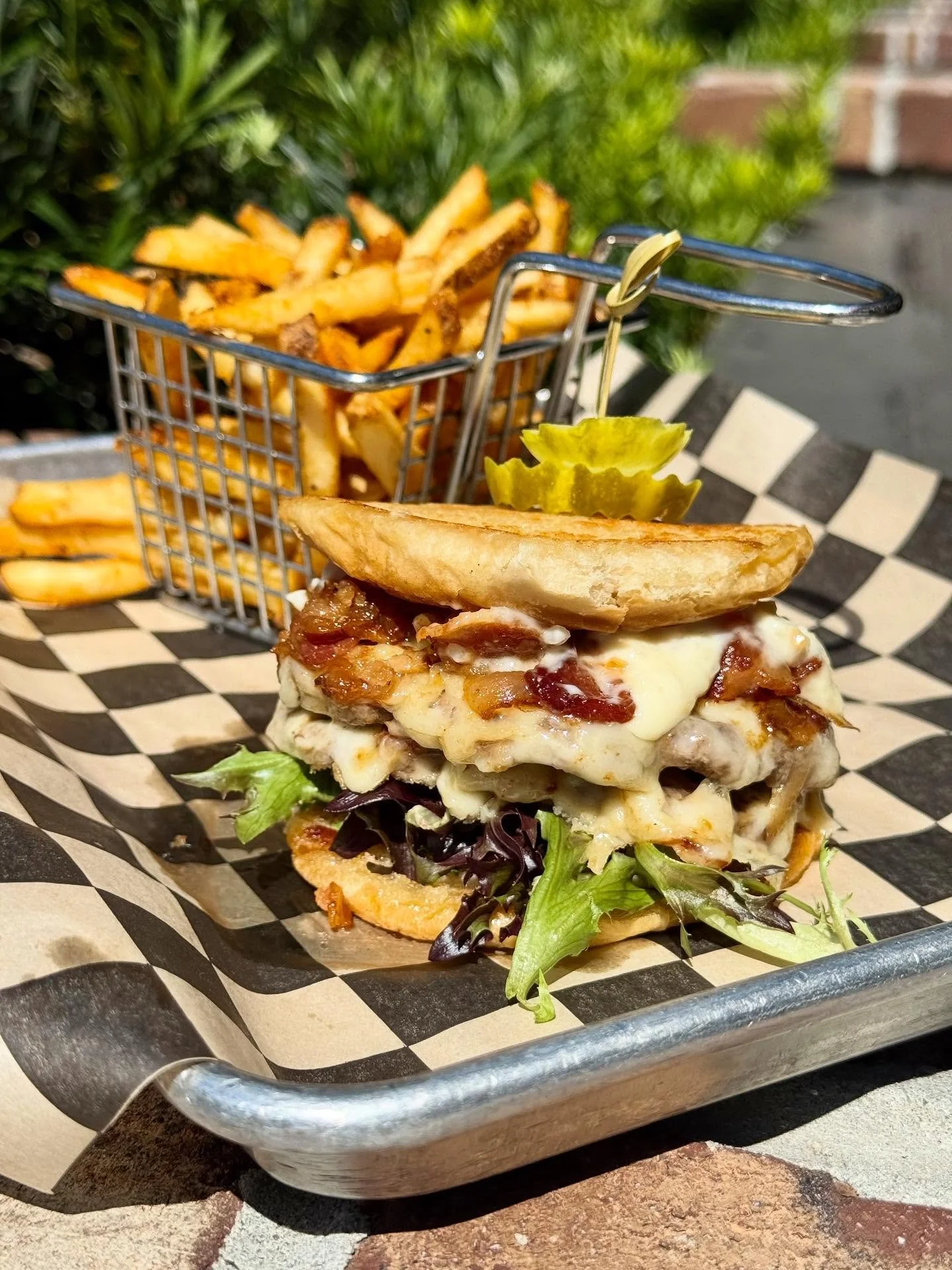 A gourmet burger with lettuce, bacon, cheese, and pickles, served with a side of French fries in a small metal basket, placed on checkered paper