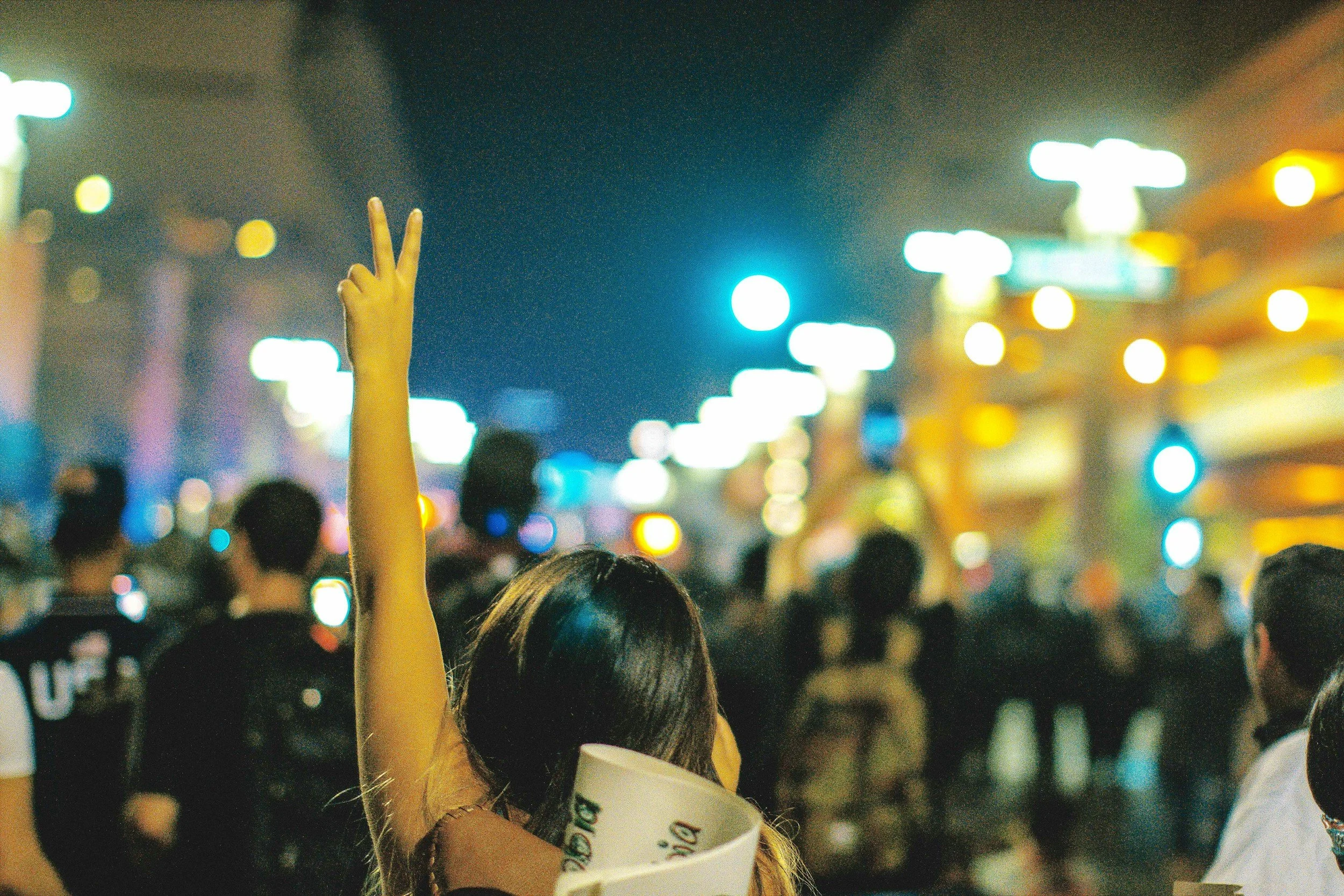 A woman seen from behind in a night crowd, one arm raised with her fingers in a peace sign.