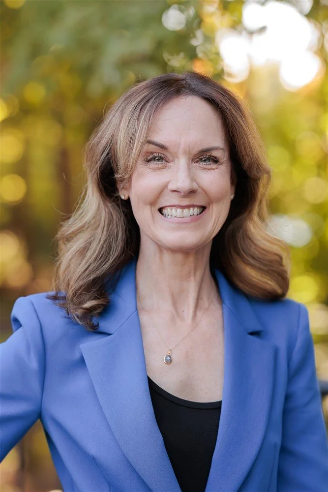 A woman with shoulder-length light brown hair, green eyes, and a bright smile, wearing a dark blazer.