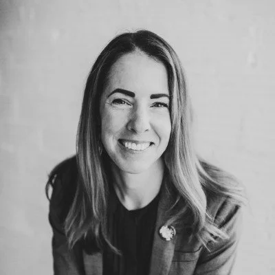 Portrait of a smiling blonde woman with blue eyes, wearing a white blazer and pink blouse, against a dark background.