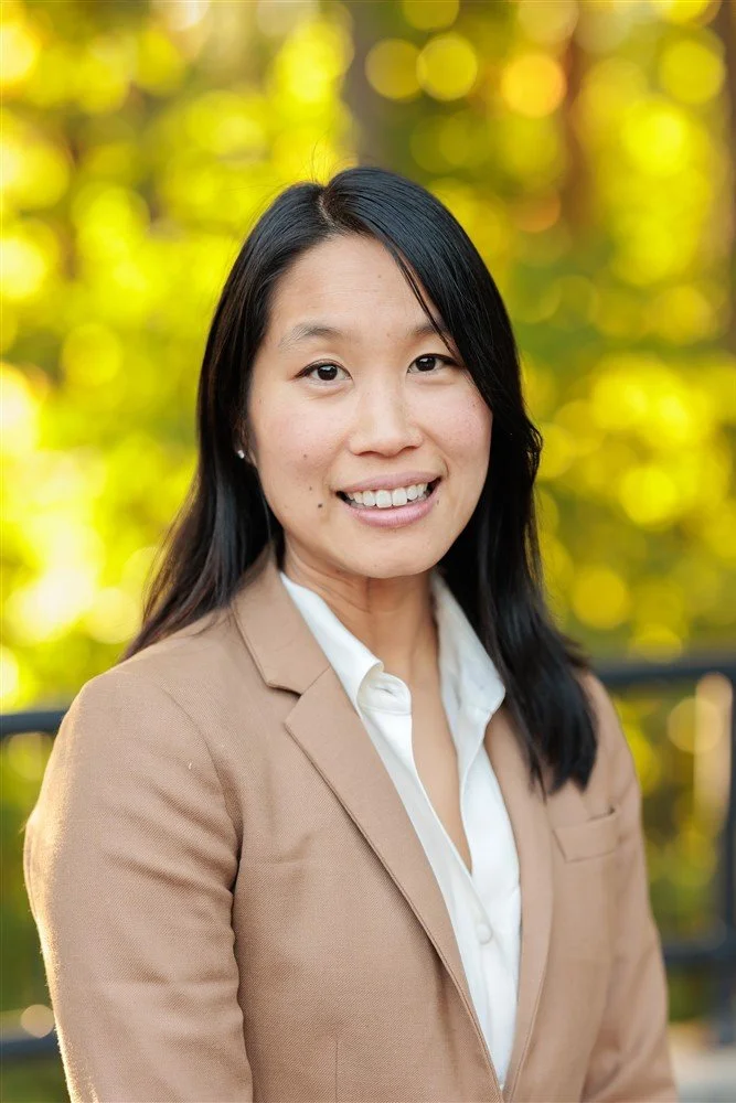 A portrait of a smiling Asian woman with dark hair and hoop earrings, wearing a beige turtleneck sweater, against a teal background.