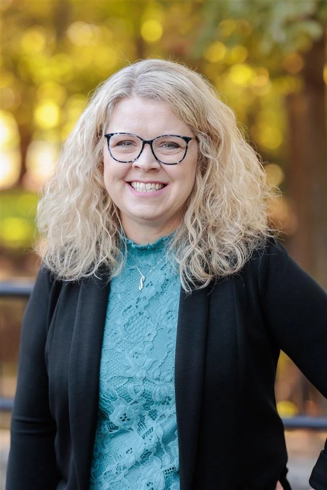 A woman with blonde curly hair, wearing glasses, a black blazer, and a blue top, smiling against a gray background.