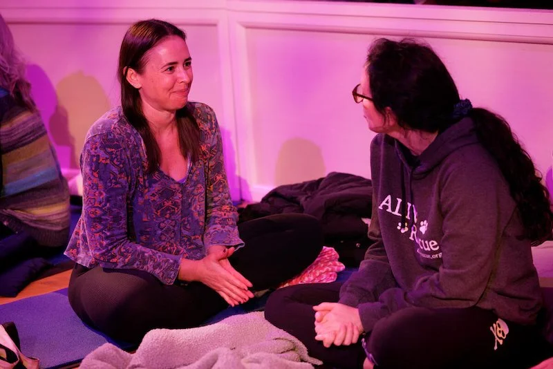 Two women sitting on the floor talking, surrounded by blankets, under pink lighting.
