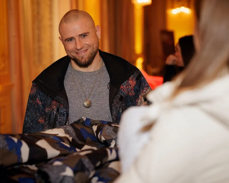 Man smiling indoors, dressed in a patterned jacket with a pendant necklace, facing a person in a white coat.