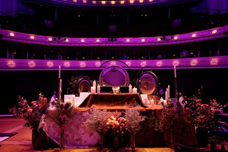 Stage setup with gongs, candles, and floral arrangements in a theater with purple lighting.