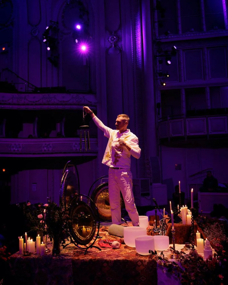 Man in white clothing stands in a dimly lit, ornate theater setting, holding a small item. Surrounded by gongs, candles, and flowers on a decorated table. Purple lighting creates a mystical ambiance.