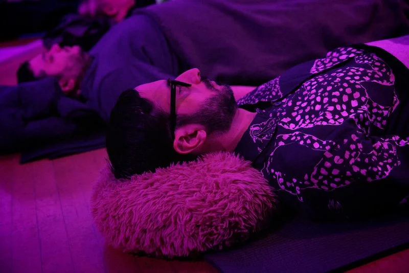 Person lying on a mat with a pillow, under pink lighting, wearing glasses and a patterned shirt.