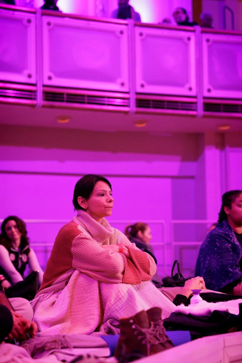 A woman sitting on a cushioned surface in a purple-lit room, wearing a cozy sweater, wrapped in a blanket, with people sitting around her.