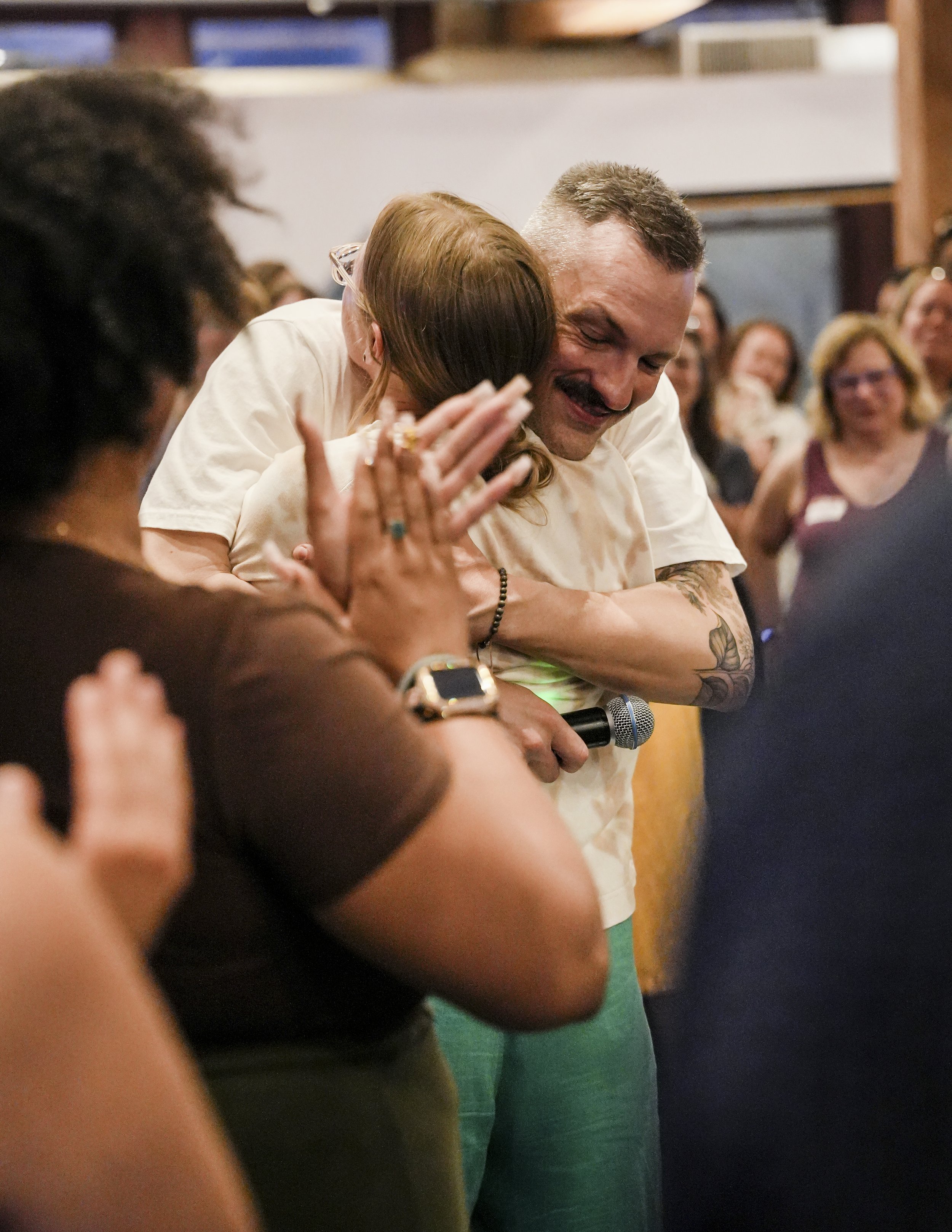 Davin Youngs singing into a microphone while hugging a woman during a gathering or event, with people watching and clapping around them.