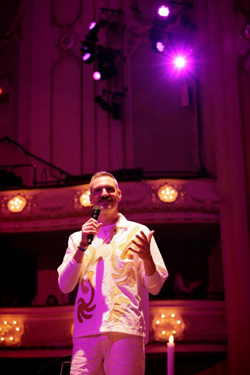 Person holding a microphone under purple stage lighting in an ornate theater.
