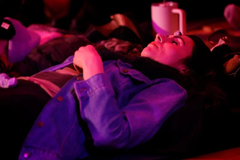 Person lying down with eyes closed, wearing a denim jacket in a dimly lit setting.