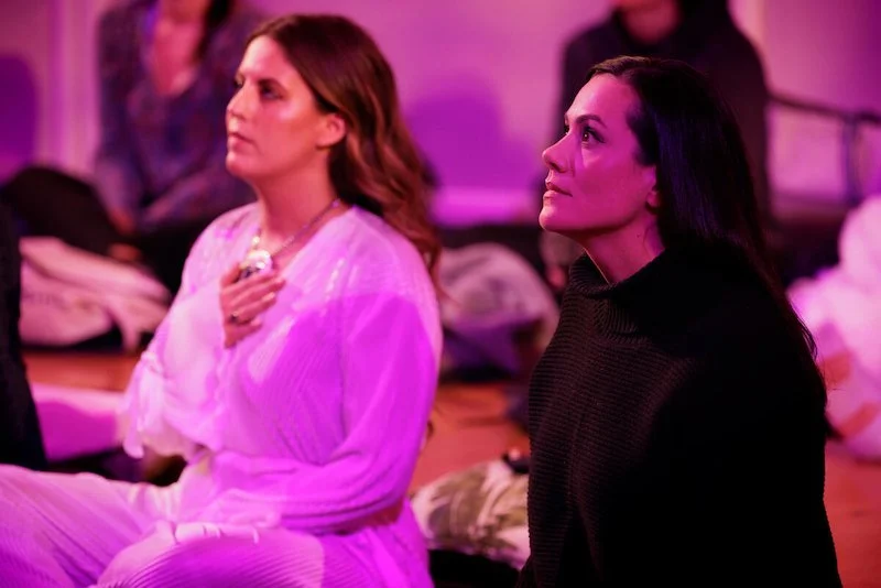 Two women sitting cross-legged in a softly lit room, one with her hand on her chest, appearing thoughtful or meditative.