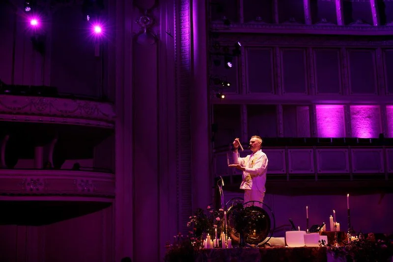Man performing on stage with purple lighting, surrounded by candles and plants, holding a small cymbal.