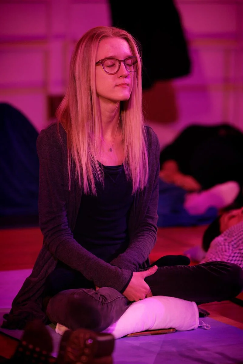 Woman meditating indoors with eyes closed, wearing glasses, seated on a cushion, surrounded by a dimly lit setting.