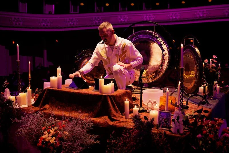Person kneeling in front of musical setup with lit candles and gongs in a dimly lit room.
