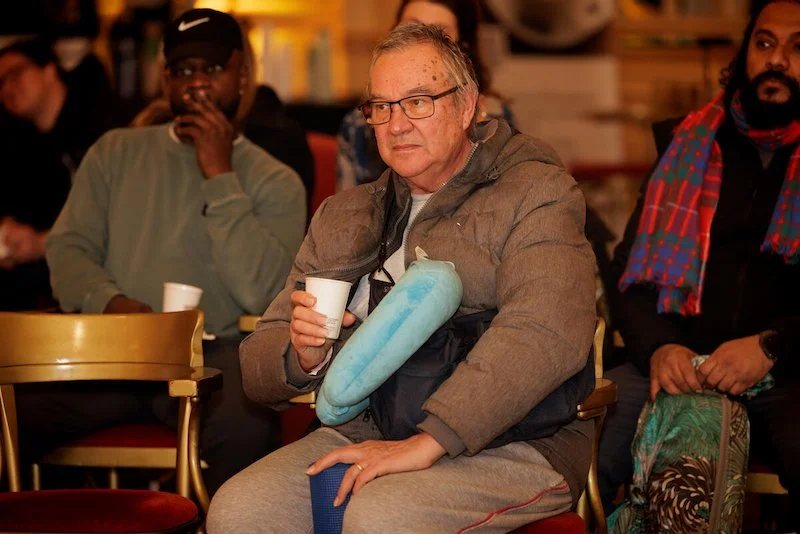 A group of people sitting in a room, listening attentively. One person in the foreground wears glasses, a puffer jacket, and holds a paper cup with a neck pillow around their neck.