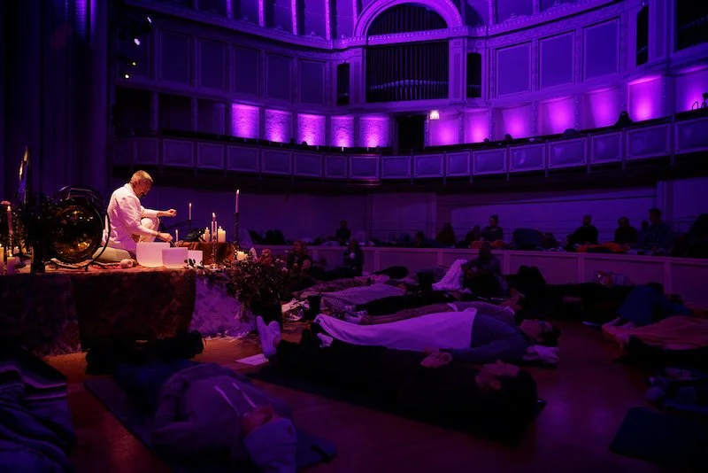 Person performing a sound bath session on stage, surrounded by people lying down with eyes closed in a dimly lit room with purple lights.