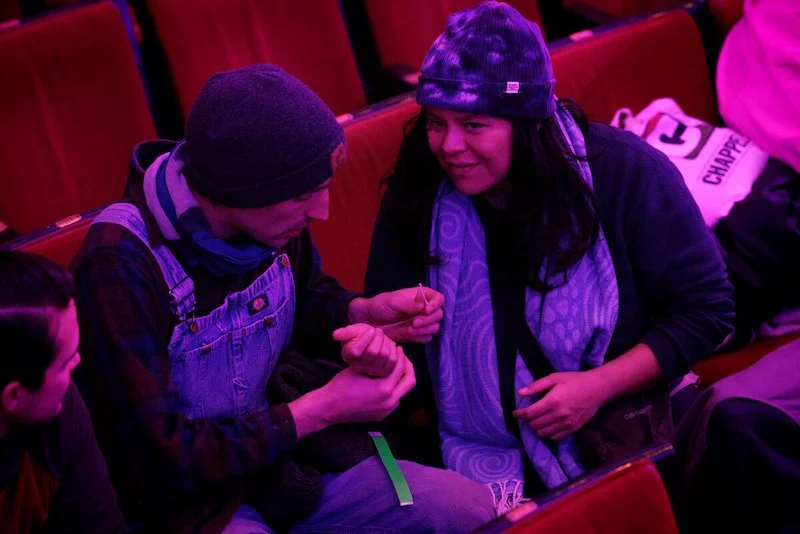 People sitting in theater seats, one wearing a beanie and overalls, engaging with another person in a winter hat and scarf.