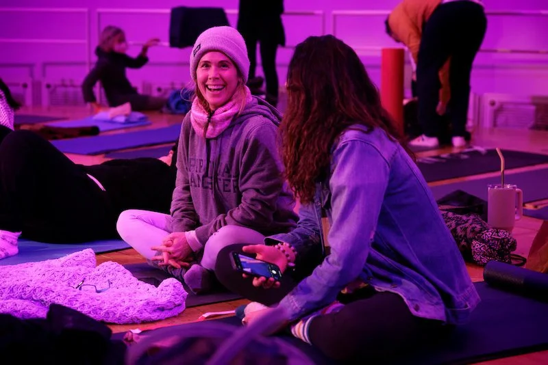 Two women sitting on yoga mats indoors, talking and smiling, with a purple ambient light.