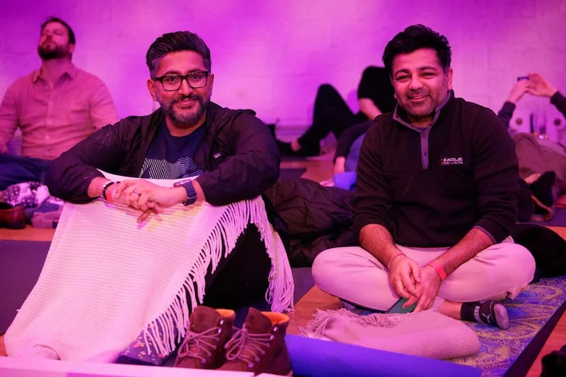 Two men sitting on yoga mats in a relaxed setting with purple lighting, wearing casual clothing, with a blanket and shoes nearby. Other people are in the background.
