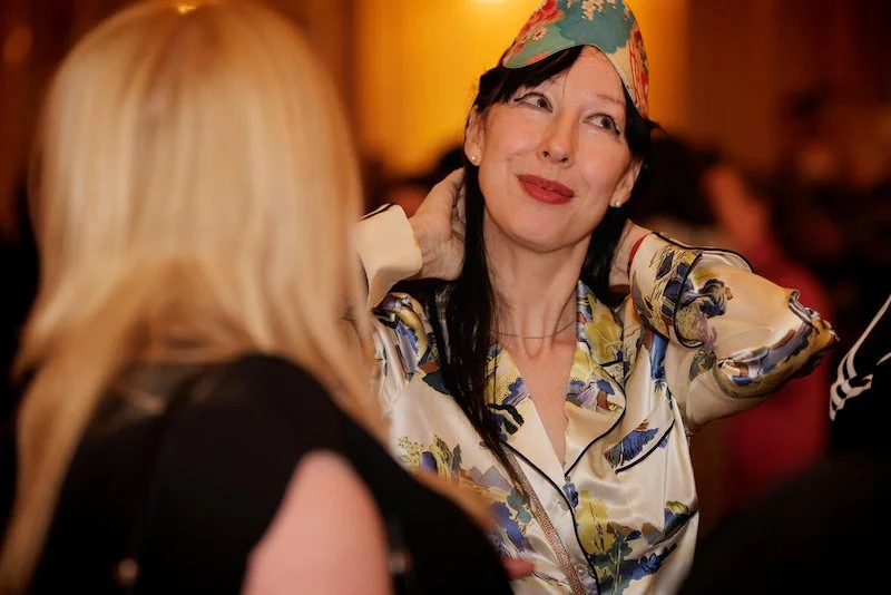 Two women talking at an event, one wearing a colorful headscarf and patterned top, blurred background.