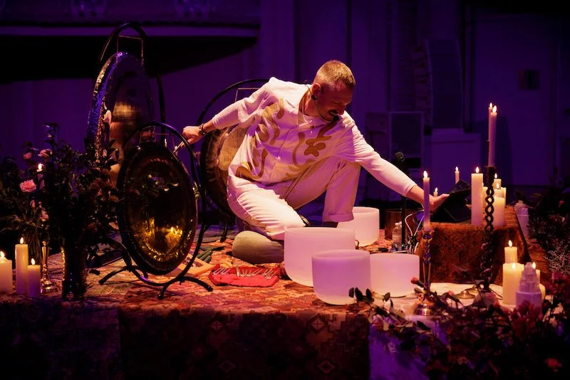 Person in white attire playing crystal singing bowls and gongs surrounded by lit candles and flowers, creating a serene atmosphere.
