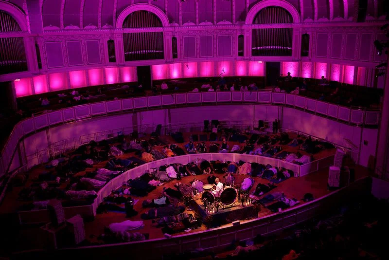 Concert hall with purple lighting and people lying on the floor gathered around a drum set.