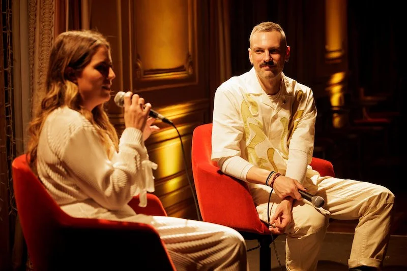 Two people sitting on red chairs on a stage holding microphones, engaging in a conversation. They are dressed in casual attire with warm lighting illuminating the background.