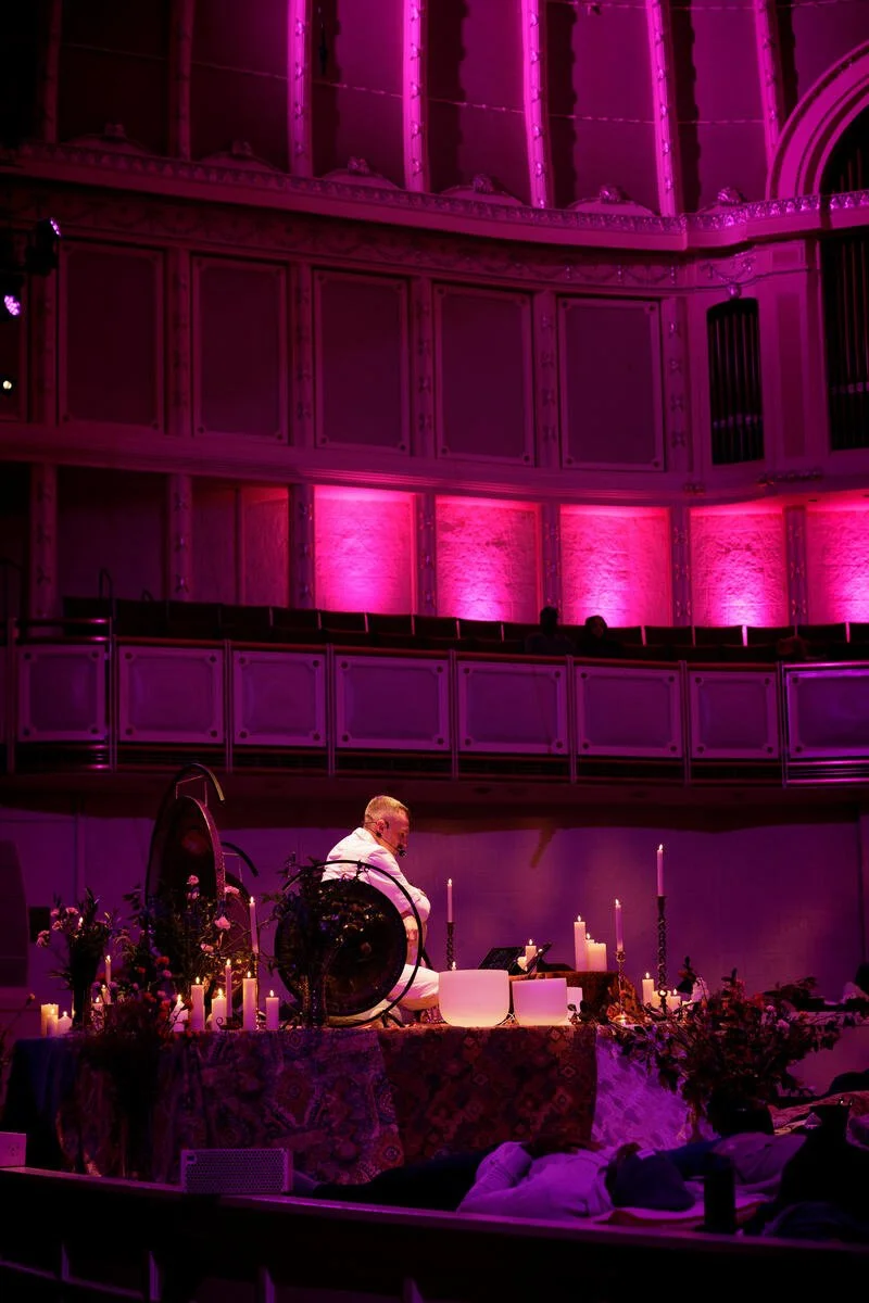 A person in a white robe is performing a meditative or ritualistic ceremony on a stage surrounded by candles and flowers. The lighting is predominantly pink, illuminating a large hall with ornate architectural details.