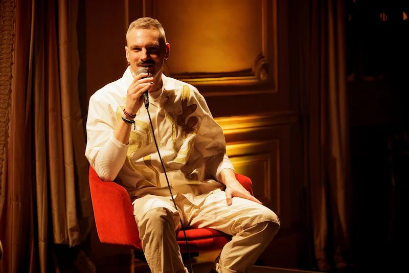 Person sitting on a red chair speaking into a microphone in a warmly lit room.
