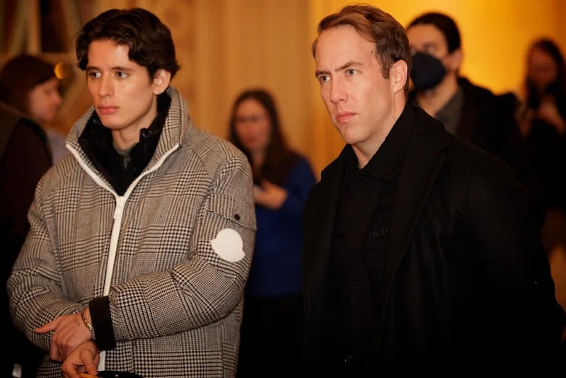 Two men standing indoors with a focused expression, one wearing a gray plaid jacket, the other in a black coat.