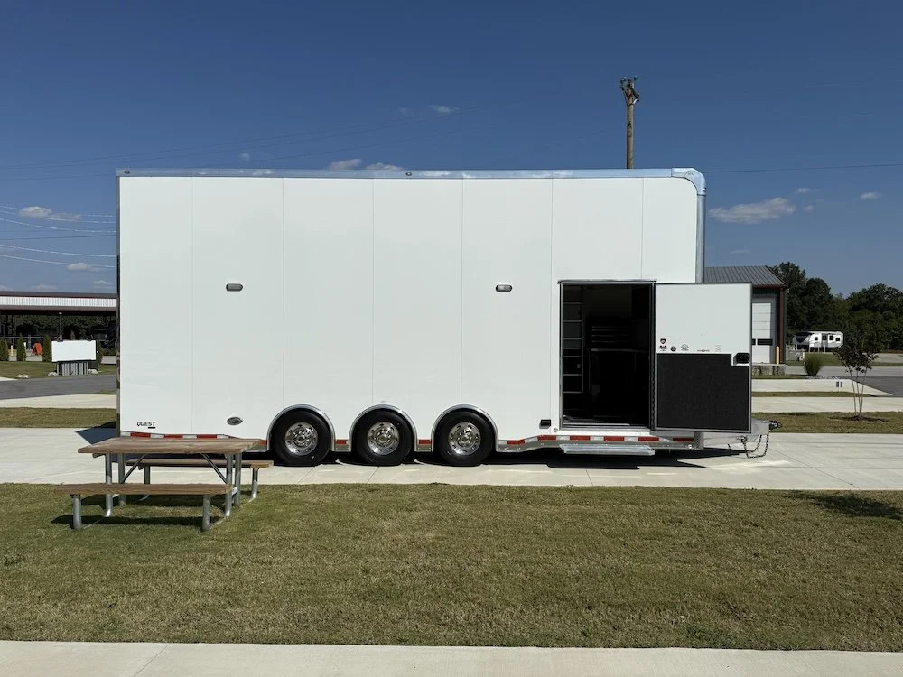A large white trailer with a side door open, parked on a paved area next to a grassy lawn with a picnic table.