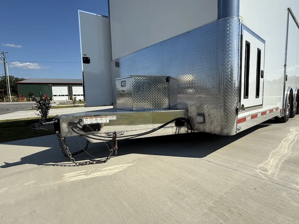 A close-up of the front of a white enclosed trailer with a diamond plate metal front. The trailer hitch and safety chains are visible, lying on the concrete ground.