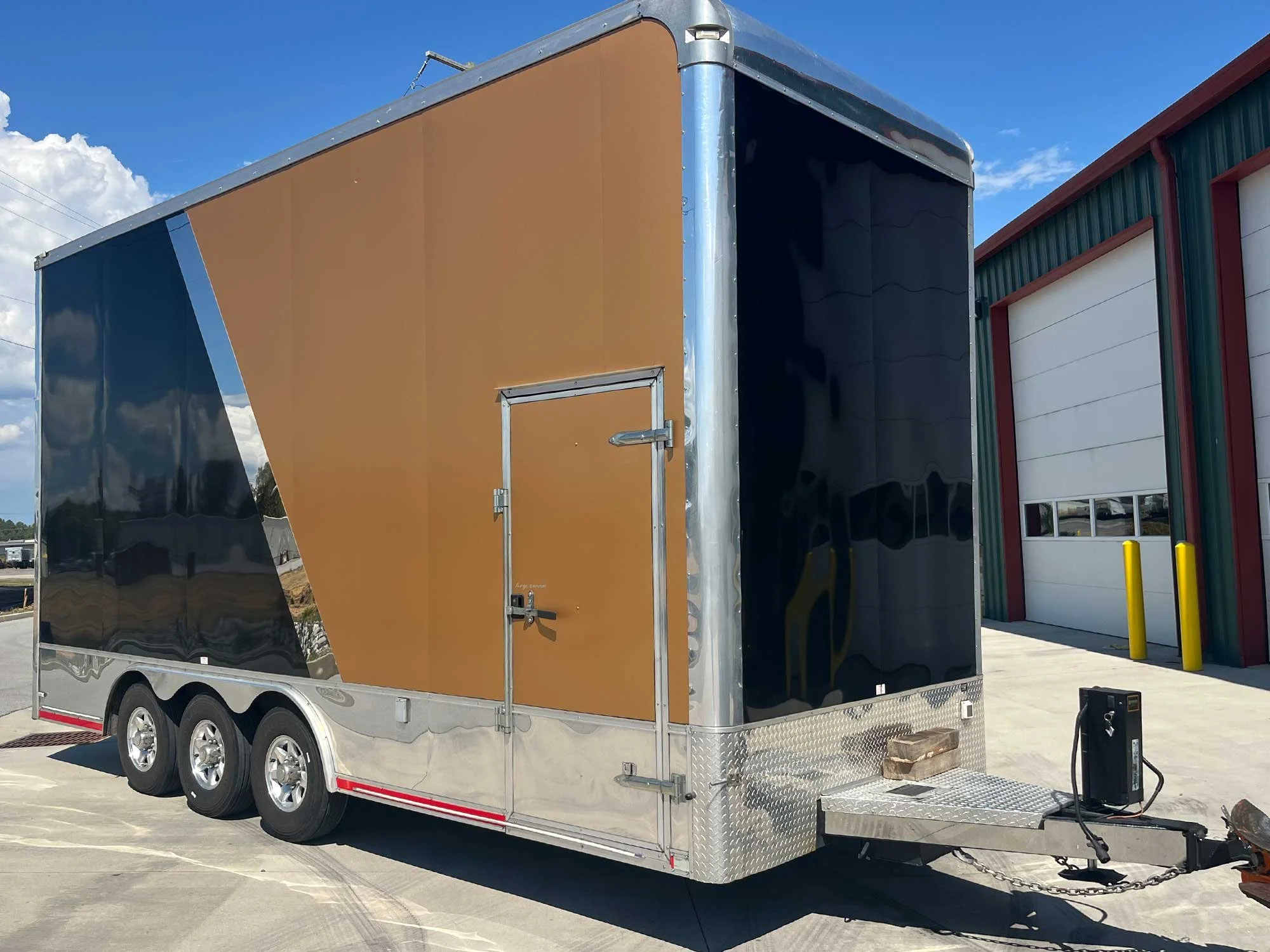 A black and gold enclosed trailer with triple axles parked on a concrete lot, with a large industrial garage with red and green siding in the background.