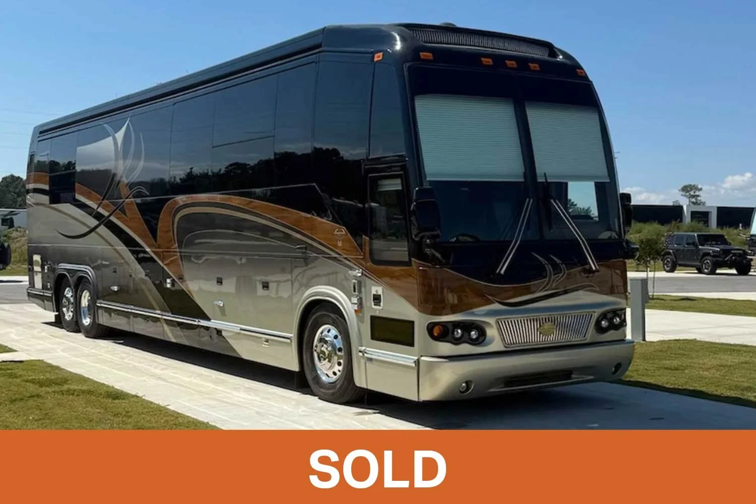 A black, brown, and beige luxury motorhome parked outdoors on a paved lot, with other RVs and vehicles in the background under a clear blue sky.