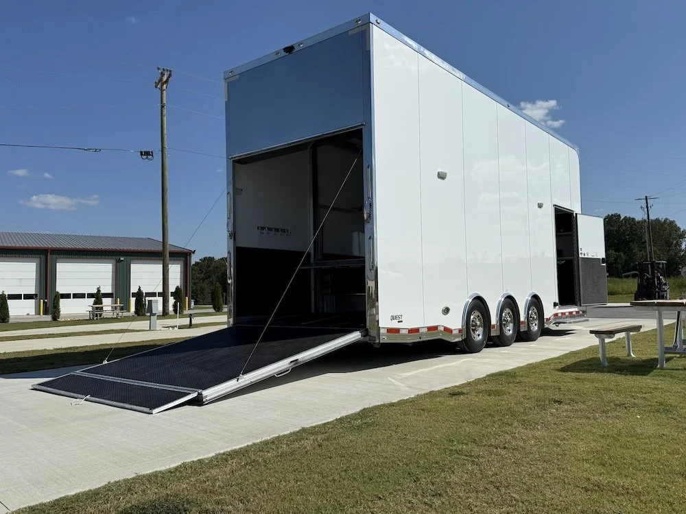 A large white trailer with a lowered ramp door on a concrete surface, with a grassy area and a building with garage doors in the background.