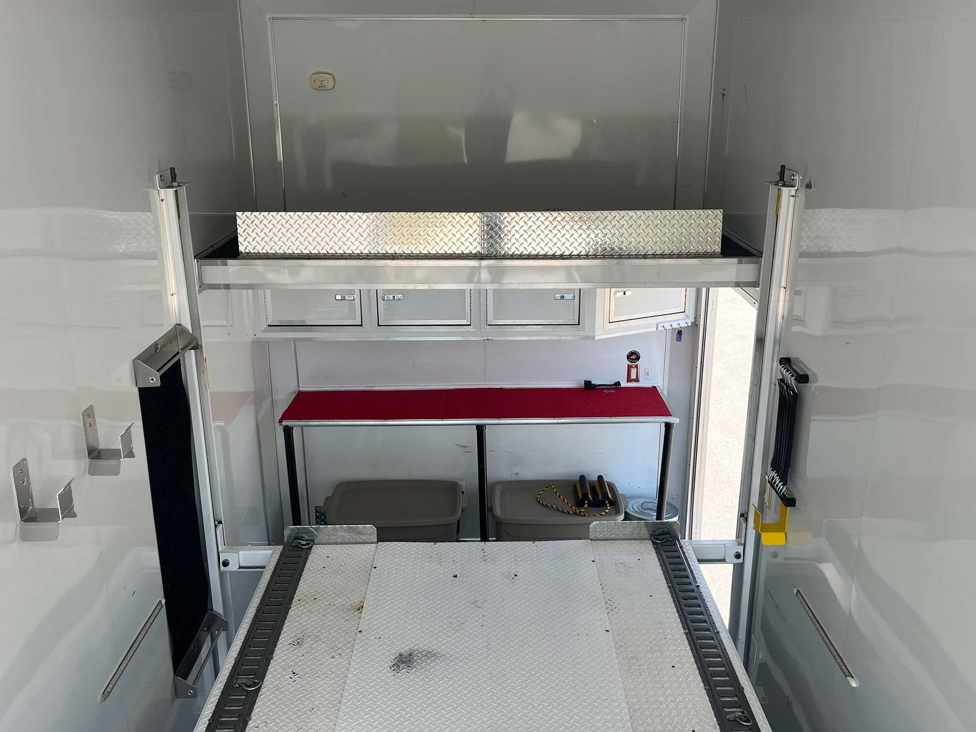 Interior of a mobile equipment trailer showing a workbench with a red mat, storage drawers above, and storage bins below. Metal racking and hooks are attached to the walls.