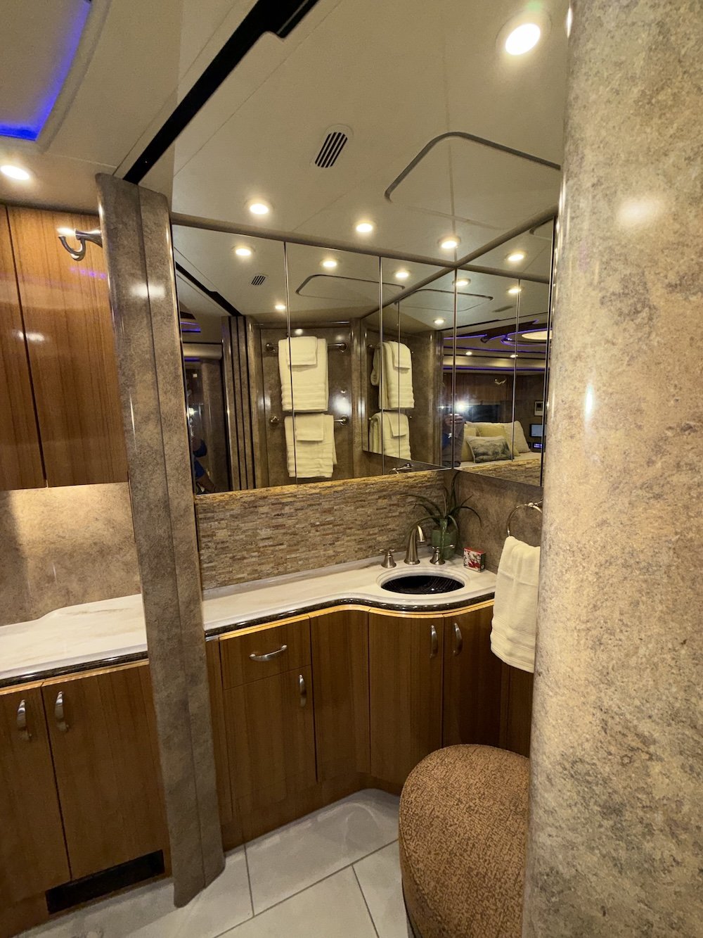 A bathroom with a large mirror, wooden cabinet with a marble countertop, a small sink with a faucet, a potted plant, and yellow towels hanging on the wall. 