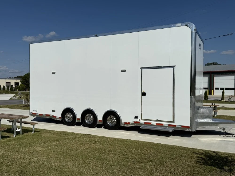A large white enclosed trailer with three axles parked on a grassy area with concrete pavers, a park bench nearby, and buildings in the background under a clear blue sky.