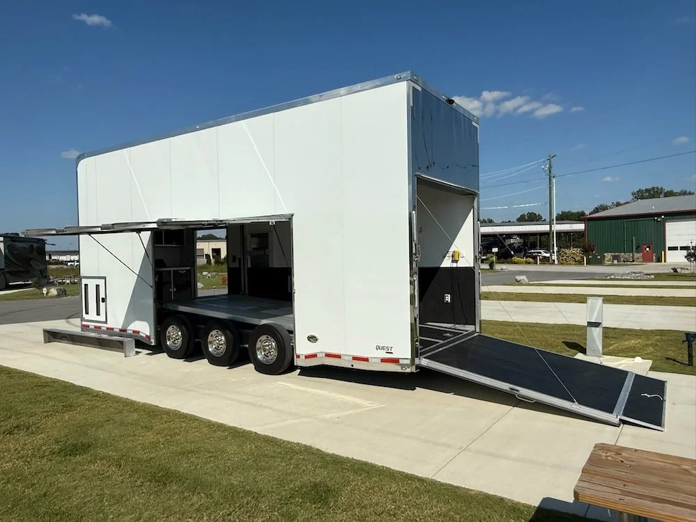 A large white trailer with three axles, open side door, and folding ramp, parked on a concrete pad in a suburban area.