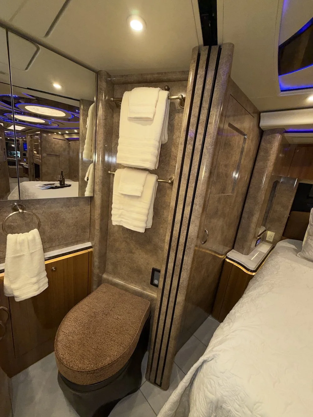 A bathroom with a bed against a wall, a mirror, towels, and a brown ottoman.