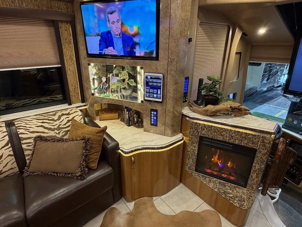 Interior of a luxurious motorhome living room featuring a mounted TV, fireplace, sofa with decorative pillows, and various electronic panels and devices.