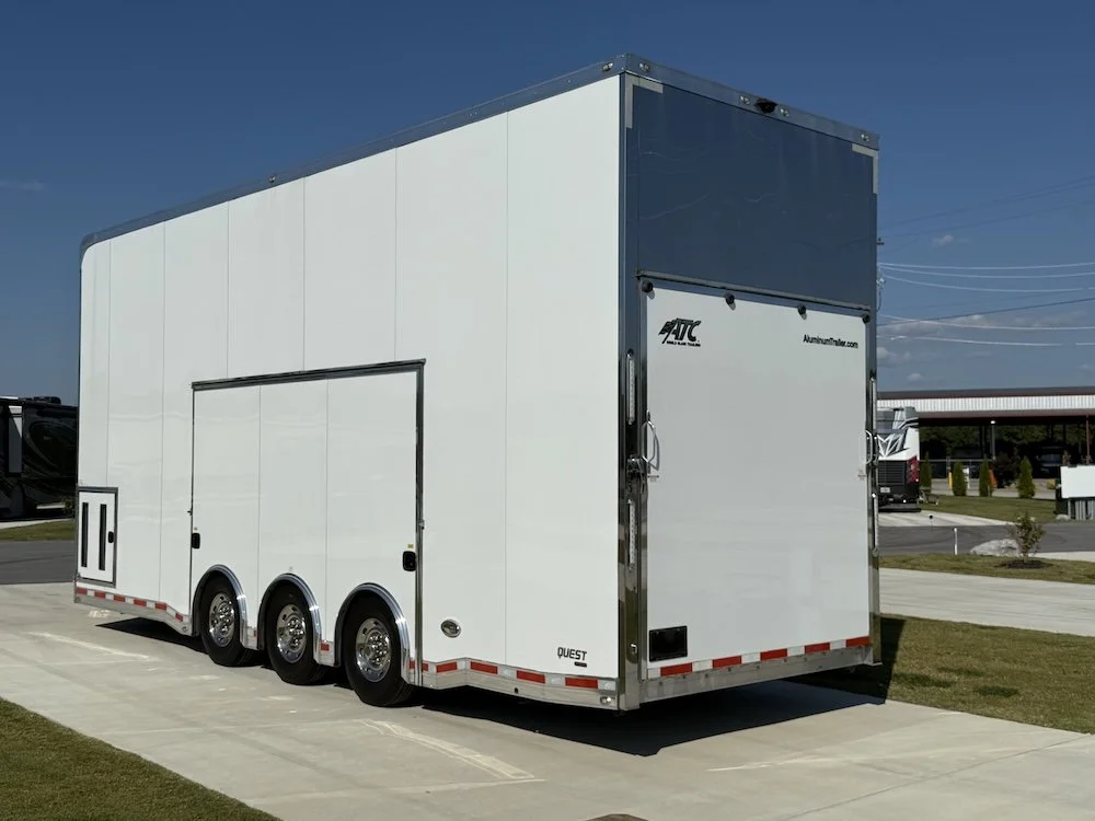 Large white enclosed trailer with triple axles parked on a paved lot with grass and other trailers in the background.