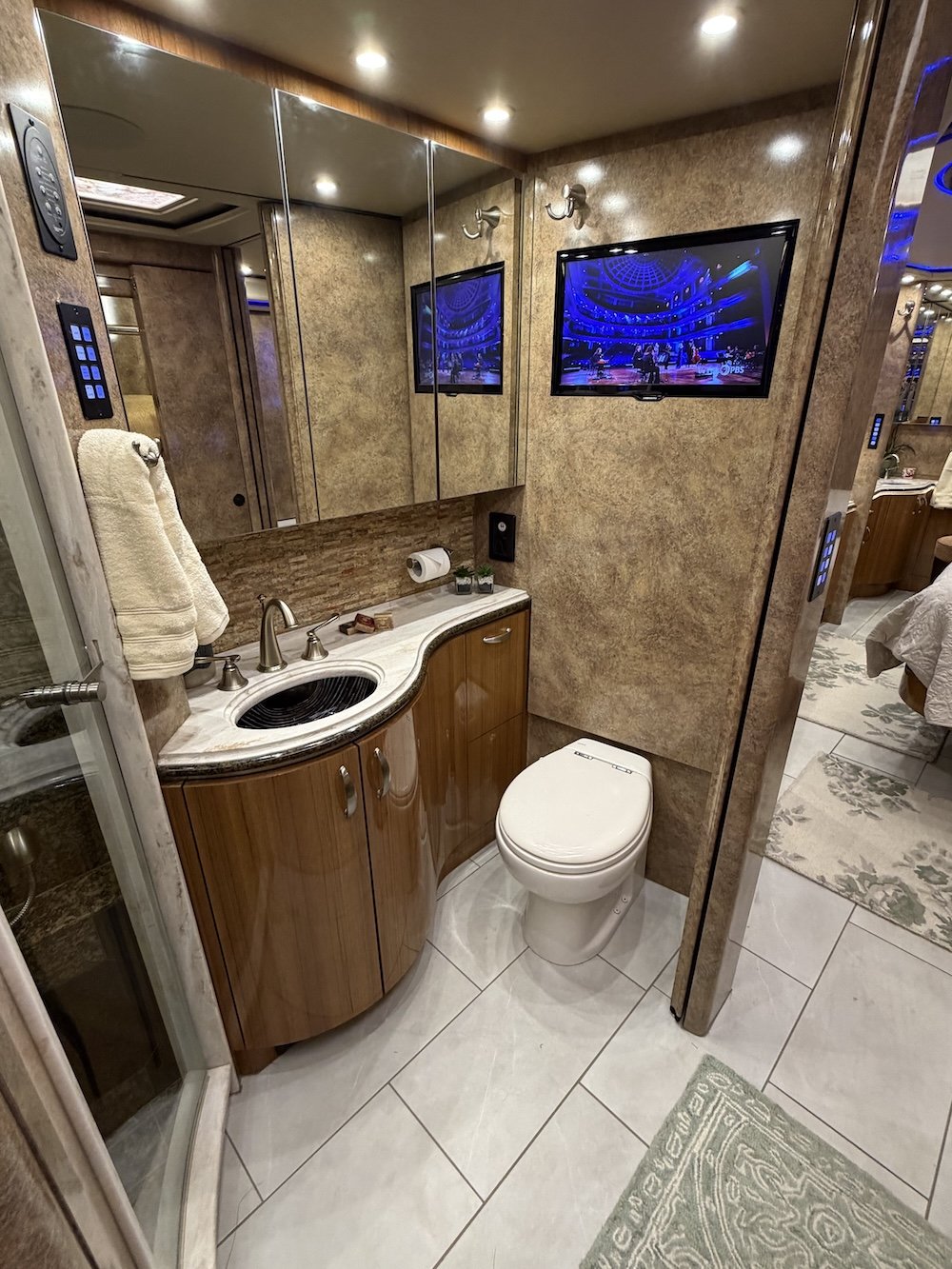 Small bathroom with a curved wooden vanity with a marble countertop, a white toilet, a sink with a faucet, wall-mounted mirror cabinets, a television on the wall showing a stage, and a towel hanging on a hook.